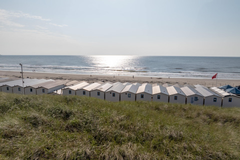 Row of beach cabins facing a tranquil sea, with grass in the foreground and sunlight on the water.