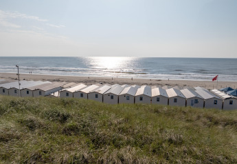 Rij strandhuisjes aan zee met gras op de voorgrond en zonlicht dat schittert op het water.