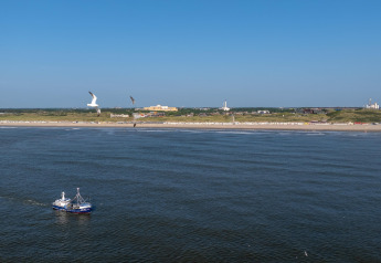 Uitzicht op zee met een vissersboot, meeuwen en een strand met gebouwen onder een blauwe lucht.