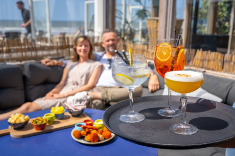 A couple relaxes at a beach house lounge with colorful cocktails and snacks served on a sunny day by the sea.
