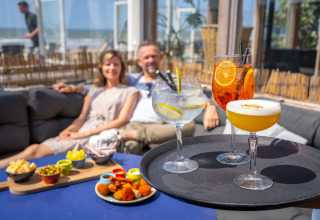 A couple relaxes at a beach house lounge with colorful cocktails and snacks served on a sunny day by the sea.