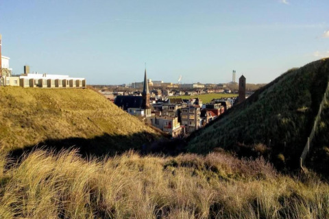 View from a beach house across grassy sand dunes, looking at a town with spires and industrial buildings beyond.