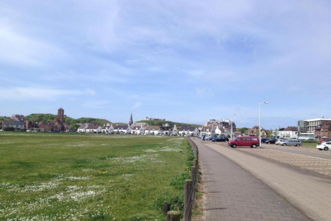 View from a beach house showing grass, a parking lot with cars, and a village under a clear blue sky.