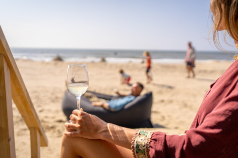 Woman relaxing with a glass of wine at a beach house while people lounge and play by the ocean.