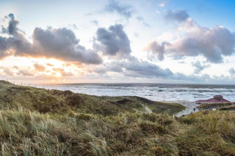 View of a beach with grassy dunes, a beach house, and dramatic clouds at sunset on the horizon.