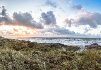 Uitzicht op een strand met begroeide duinen, een strandhuis en indrukwekkende wolken bij zonsondergang.