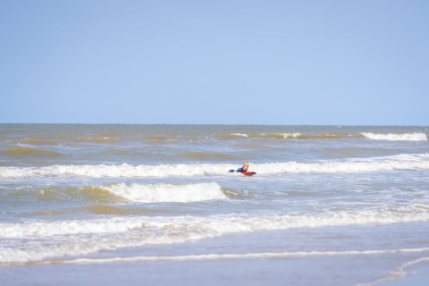 A child playing on a surfboard in the sea near the beach with gentle waves and a clear blue sky.