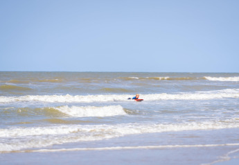 Een kind speelt op een surfplank in zee bij het strand, met zachte golven en een heldere blauwe lucht.
