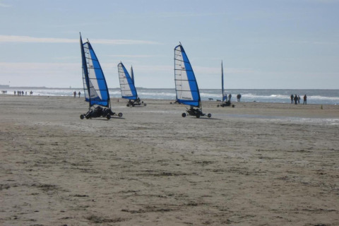 Land yachts with blue sails race along a sandy beach with people walking by the shoreline on a sunny day.