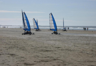 Land yachts with blue sails race along a sandy beach with people walking by the shoreline on a sunny day.