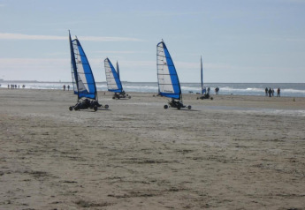 Strandzeilers met blauwe zeilen rijden over het strand terwijl mensen langs de kustlijn wandelen in de zon.