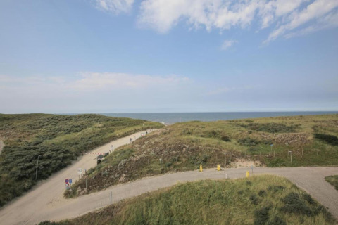 View from a beach house showing sandy dunes, winding paths, and the sea in the background under blue sky.