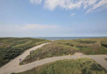 Uitzicht vanuit een strandhuis met zandduinen, kronkelende paden en de zee op de achtergrond onder blauwe lucht.