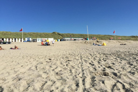 Beach scene with people, small beach houses, and red flags under a clear blue sky on a sunny day.