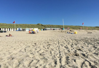 Strandscène met mensen, kleine strandhuisjes en rode vlaggen onder een heldere blauwe lucht op een zonnige dag.
