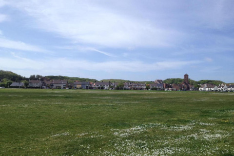 Photo of a green open field by a beach house row, with a tower building in the distance, blue sky above.