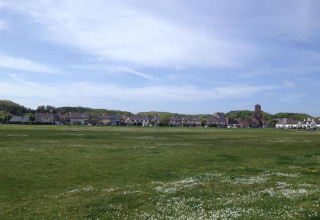 Photo of a green open field by a beach house row, with a tower building in the distance, blue sky above.