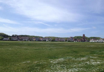 Foto van een groen veld bij een rij strandhuizen met een torengebouw, onder een heldere blauwe lucht.