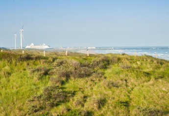 Uitzicht vanuit een strandhuis over groene duinen met windmolens, een cruiseschip en de zee in de verte.