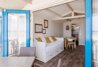 Licht strandhuisinterieur met witte bank, gele kussens, houten meubels en zeezicht door blauwe deur.