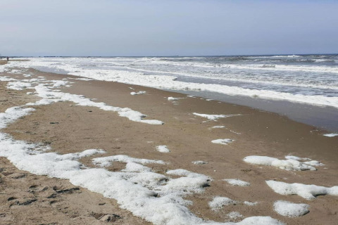 Beach scene with brown sand, sea foam scattered along the shore, and waves under a lightly clouded sky.