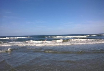 Uitzicht op de zee vanaf een strandhuis, met zachte golven en een heldere blauwe lucht erboven.