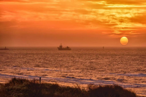 Sunset over the sea seen from a beach house, with a ship on the horizon and an orange sky above.