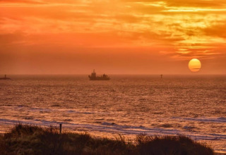 Sunset over the sea seen from a beach house, with a ship on the horizon and an orange sky above.