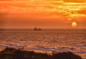 Zonsondergang boven zee gezien vanaf een strandhuis, met een schip aan de horizon en oranje lucht.