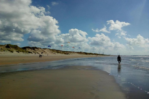 Beach scene with dunes, people walking and colorful kites in the sky beneath scattered clouds by the shore.