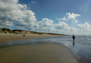 Beach scene with dunes, people walking and colorful kites in the sky beneath scattered clouds by the shore.