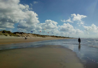 Strandtafereel met duinen, wandelende mensen en kleurrijke vliegers onder een bewolkte blauwe lucht.