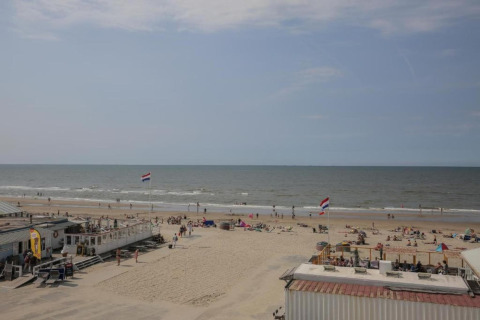 View of a beach house, people relaxing on the sand, and two Dutch flags waving by the shoreline.