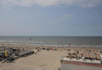 Uitzicht op een strandpaviljoen, mensen ontspannen op het strand en twee wapperende Nederlandse vlaggen.
