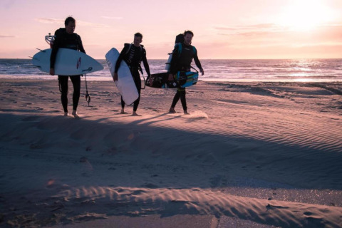 Three people in wetsuits carrying surfboards walk on the beach at sunset at Wijk aan Zee in the Netherlands.