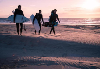 Three people in wetsuits carrying surfboards walk on the beach at sunset at Wijk aan Zee in the Netherlands.