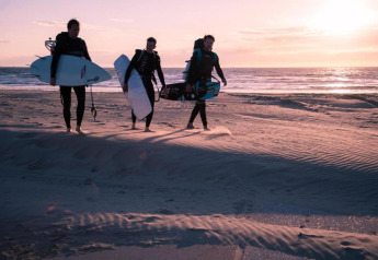 Drie mensen in wetsuits dragen surfplanken bij zonsondergang op het strand van Wijk aan Zee, Nederland.