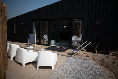 Terraza en Sea Lodges Zandvoort 3, Países Bajos, con sillas blancas y tumbonas frente a una casa negra.