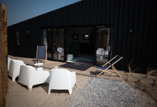 Terraza en Sea Lodges Zandvoort 3, Países Bajos, con sillas blancas y tumbonas frente a una casa negra.