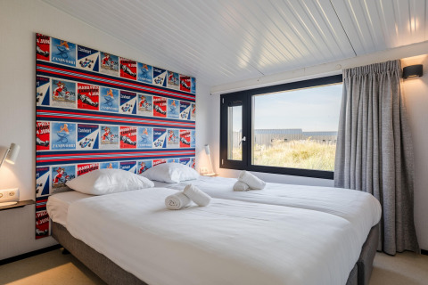 Dormitorio luminoso en casa de playa con cama doble, gran ventana y cabecero decorado con carteles coloridos.