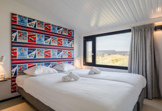 Dormitorio luminoso en casa de playa con cama doble, gran ventana y cabecero decorado con carteles coloridos.