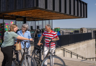 Tres personas conversan junto a bicicletas frente a una casa de playa moderna y soleada.