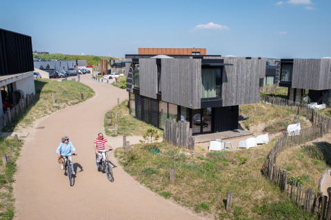 Dos personas montan bicicletas por un camino junto a casas de playa modernas en un día soleado y claro.