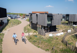 Dos personas montan bicicletas por un camino junto a casas de playa modernas en un día soleado y claro.