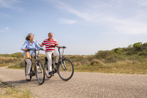 Pareja mayor pasea en bicicleta por un camino soleado junto a dunas cerca del mar, disfrutando al aire libre.