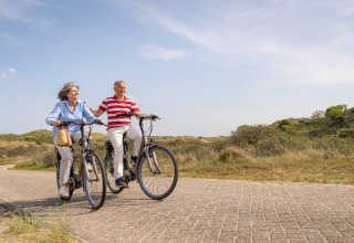 Pareja mayor pasea en bicicleta por un camino soleado junto a dunas cerca del mar, disfrutando al aire libre.