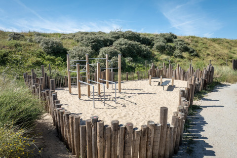Zona de ejercicios al aire libre en una casa de playa con postes de madera, barras y suelo de arena.