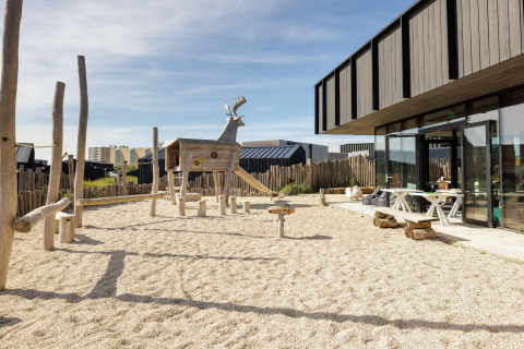 Parque infantil con animal de madera y arena junto a una casa de playa moderna con área de descanso exterior.