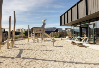 Parque infantil con animal de madera y arena junto a una casa de playa moderna con área de descanso exterior.