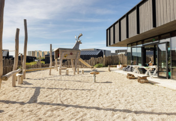 Speelplein met houten dier en zand naast een modern strandhuis met openlucht zithoek buiten.